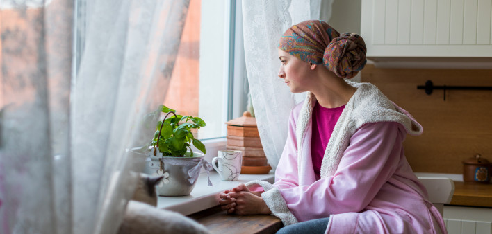 Young adult female cancer patient wearing headscarf and bathrobe sitting in the kitchen with her pet cat, looking out window.