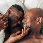 two black gay men holding hands and smiling in bed