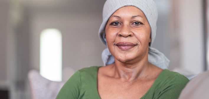 A mature black woman with cancer is wearing a scarf on her head. She is sitting at home in her living room. She is smiling directly at the camera. She is full of gratitude and hope for recovery.