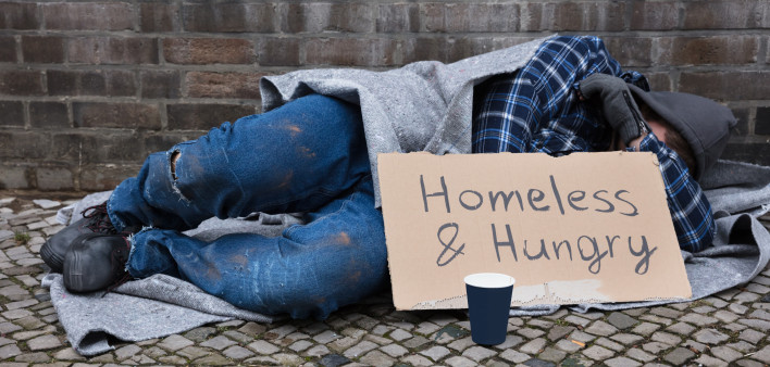 Male Beggar Lying On Street With Homeless And Hungry Text On Cardboard