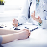 Doctor and female patient sitting at the desk and talking in clinic near window. Medicine and health care