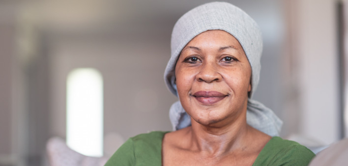 A mature black woman with cancer is wearing a scarf on her head. She is sitting at home in her living room. She is smiling directly at the camera. She is full of gratitude and hope for recovery.