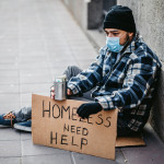 Homeless young man sitting on sidewalk and begging alone. He is wearing protective face mask and holding out a can and begging for money. His head is bowed in shame and tiredness and hunger.