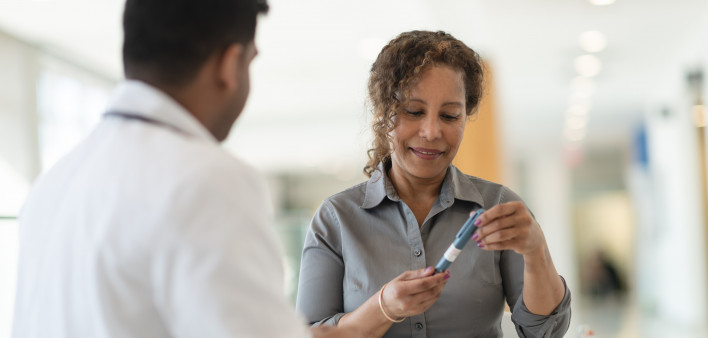 A male Indian doctor meets with a female patient. The woman is a mature adult of African descent. The two people are seated at a table. The patient is diabetic and is holding an insulin pen.