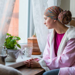 Young adult female cancer patient wearing headscarf and bathrobe sitting in the kitchen with her pet cat, looking out window.