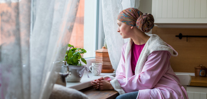 Young adult female cancer patient wearing headscarf and bathrobe sitting in the kitchen with her pet cat, looking out window.