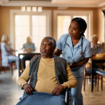 African American senior in wheelchair and young nurse looking through the window at residential care home.