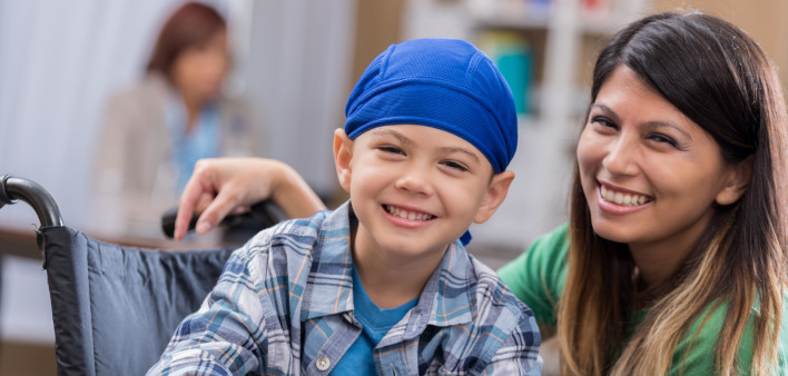 Mid adult mom and her elementary age son wait in waiting room in doctor's office or hospital. The boy is wearing a blue cap and is sitting in a wheelchair.