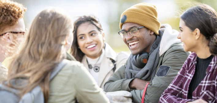 Five young adults are sitting at a picnic table outside on a fall day. They are smiling and chatting