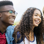 A girl of African American descent smiles with laughter while sitting in a row of teens with the arms around each other's shoulders. They are laughing too.