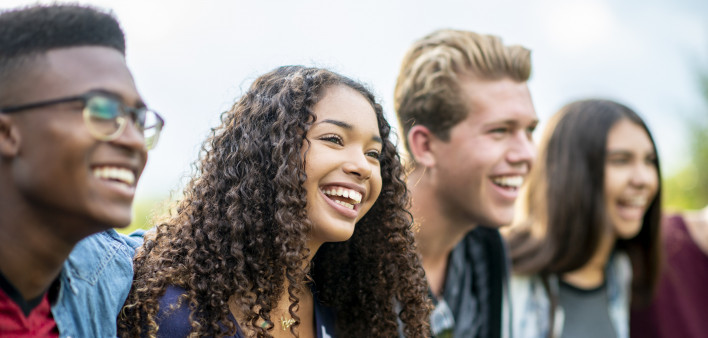 A girl of African American descent smiles with laughter while sitting in a row of teens with the arms around each other's shoulders. They are laughing too.
