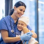 Nurse Hugging Young Cancer Patient stock photo A young Latin female nurse hugs her cancer patient as they enjoy a brief moment together. The young girl is smiling and happy. The nurse is wearing blue scrubs with a stethoscope around her neck, while the yo