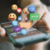 Hand holding a smart phone with various emojis emerging off the screen.