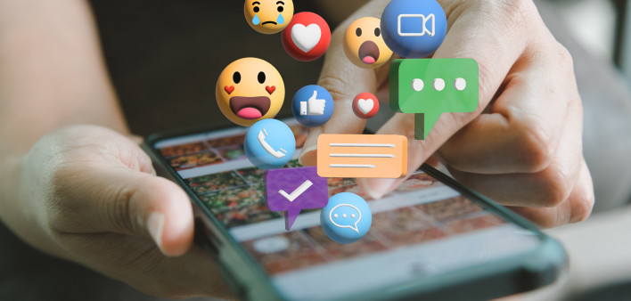 Hand holding a smart phone with various emojis emerging off the screen.