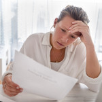 Woman looking worried holding paperwork at home. She is reading a financial bill or a letter with bad news. She looks very stressed and upset.