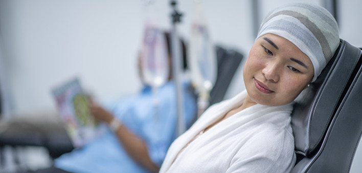 A woman wearing a head scarf lies on a hospital bed and looks to the side in contemplation. She is wearing a head scarf and a hospital gown and there is a IV drip next to her.
