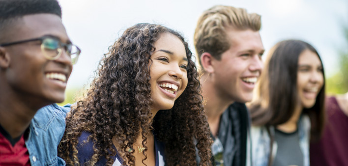 A girl of African American descent smiles with laughter while sitting in a row of teens with the arms around each other's shoulders. They are laughing too.