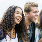 A girl of African American descent smiles with laughter while sitting in a row of teens with the arms around each other's shoulders. They are laughing too.
