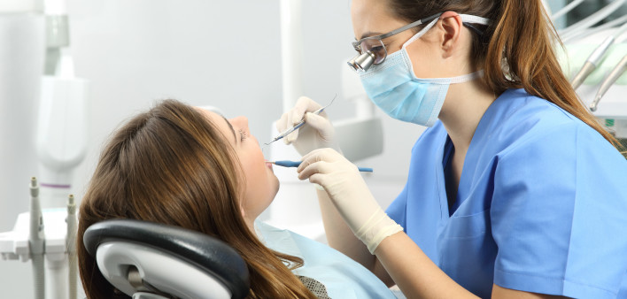 Dentist wearing eyeglasses gloves and mask examining a patient teeth with a dental probe and a mirror in a clinic box with equipment in the background