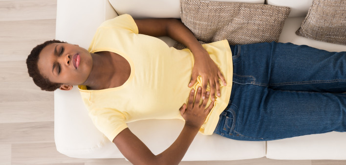 Young African Woman Lying On Sofa Suffering From Stomach Ache