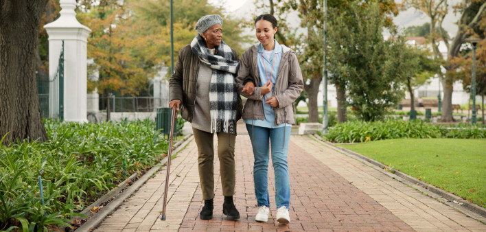 Help, cane and nurse with old woman in park for relax, support and person with a disability. Elderly care, conversation and healthcare with caregiver and patient in nature for medical rehabilitation