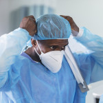 Two multiracial healthcare workers working in a hospital ward, putting on full PPE. They are wearing surgical caps and gowns and adjusting their N95 face masks.