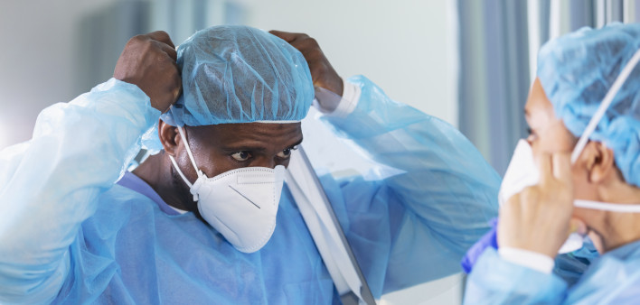 Two multiracial healthcare workers working in a hospital ward, putting on full PPE. They are wearing surgical caps and gowns and adjusting their N95 face masks.