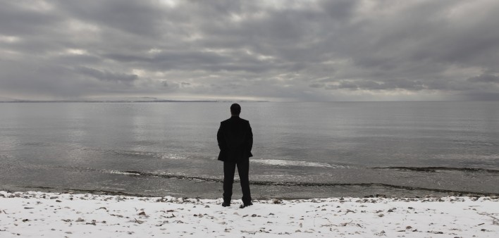 A man stands alone facing the ocean with a cloudy sky above