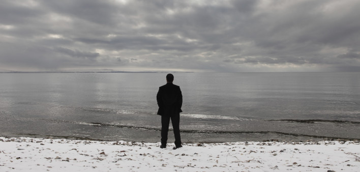 A man stands alone facing the ocean with a cloudy sky above