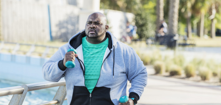 A mature African-American man in his 40s with a large build, exercising outdoors in a city park. He is wearing a fitness tracker, jogging or power walking with hand weights.