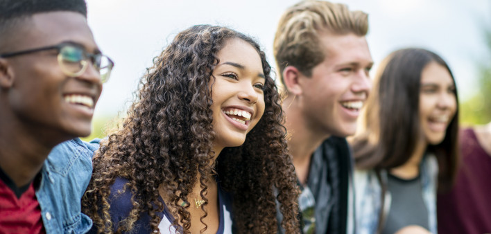 A girl of African American descent smiles with laughter while sitting in a row of teens with the arms around each other's shoulders. They are laughing too.