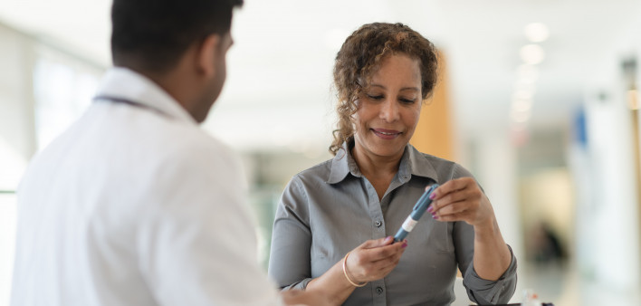 A male Indian doctor meets with a female patient. The woman is a mature adult of African descent. The two people are seated at a table. The patient is diabetic and is holding an insulin pen.