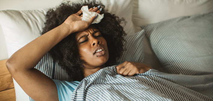 Young African-American woman feeling sick at home. Lying in bed, drinking tea and medicine. Daytime.
