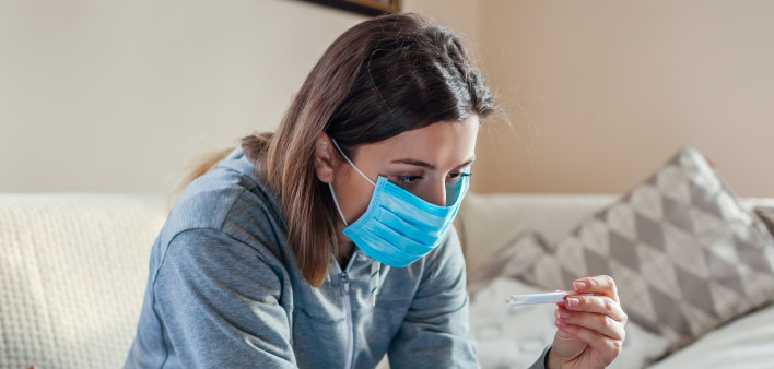 Ill infected woman checking thermometer having fever. Girl wearing protective mask while having cold, flu taking pills at home. Healthcare