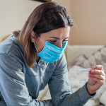 Ill infected woman checking thermometer having fever. Girl wearing protective mask while having cold, flu taking pills at home. Healthcare