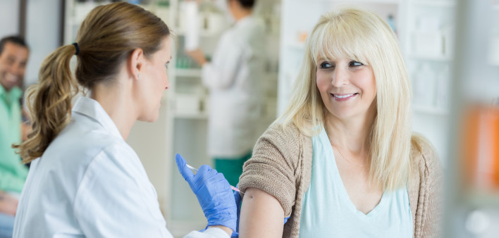pharmacist wears a lab coat and medical gloves as she pushes a needle into the arm of an apprehensive, but smiling mid adult female patient. The patient is looking at the pharmacist...