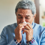 sickly senior man blowing his nose with a tissue at home