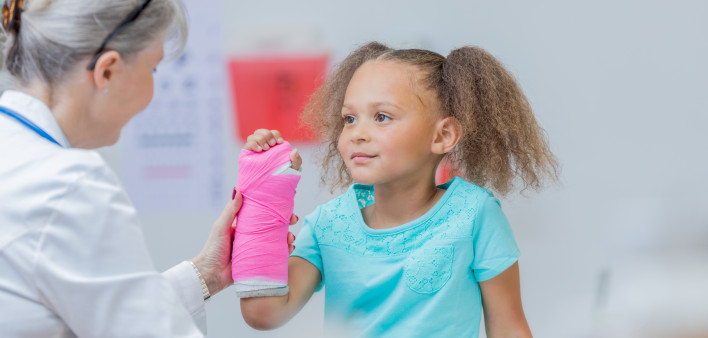 Pediatric orthopedic doctor examines an elementary age mixed race girl's broken arm. The girl has a pink cast on her arm.