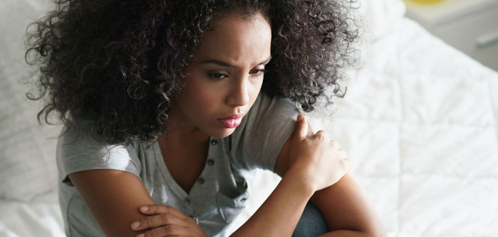Lonely young latina woman sitting on bed. Depressed hispanic girl at home, looking away with sad expression.
