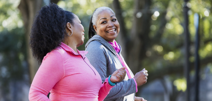 Two African-American women exercising together in the city, jogging or power walking, laughing and conversing. Buildings and trees are out of focus in the background. The one in pink is in her 60s...