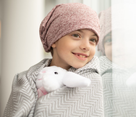 A happy elementary age girl with cancer is wearing a pink scarf on her head.