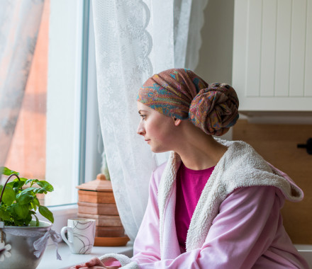 Young adult female cancer patient wearing headscarf and bathrobe sitting in the kitchen with her pet cat, looking out window.