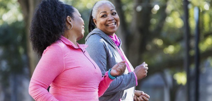 Two African-American women exercising together in the city, jogging or power walking, laughing and conversing. Buildings and trees are out of focus in the background. The one in pink is in her 60s...