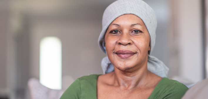 A mature black woman with cancer is wearing a scarf on her head. She is sitting at home in her living room. She is smiling directly at the camera. She is full of gratitude and hope for recovery.