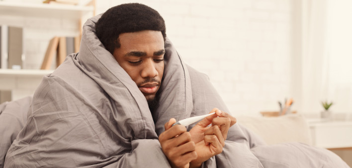 Ill african-american man covered with blanket holding thermometer, having fever