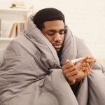 Ill african-american man covered with blanket holding thermometer, having fever
