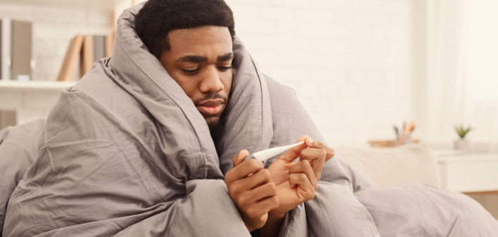 Ill african-american man covered with blanket holding thermometer, having fever
