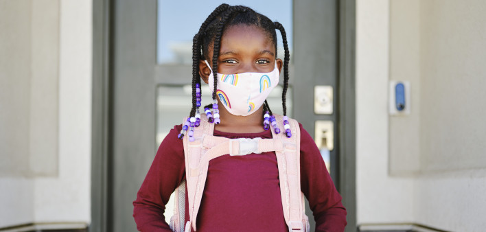 An elementary aged school student leaving her home to go back to school, wearing a mask for protection against infectious disease.