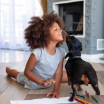 Funny little African American girl lying on floor coloring picture having fun with dog,