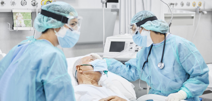 Female doctor talking with patient along coworker in ICU. Man is lying on bed amidst essential workers. Healthcare workers are in protective workwear.